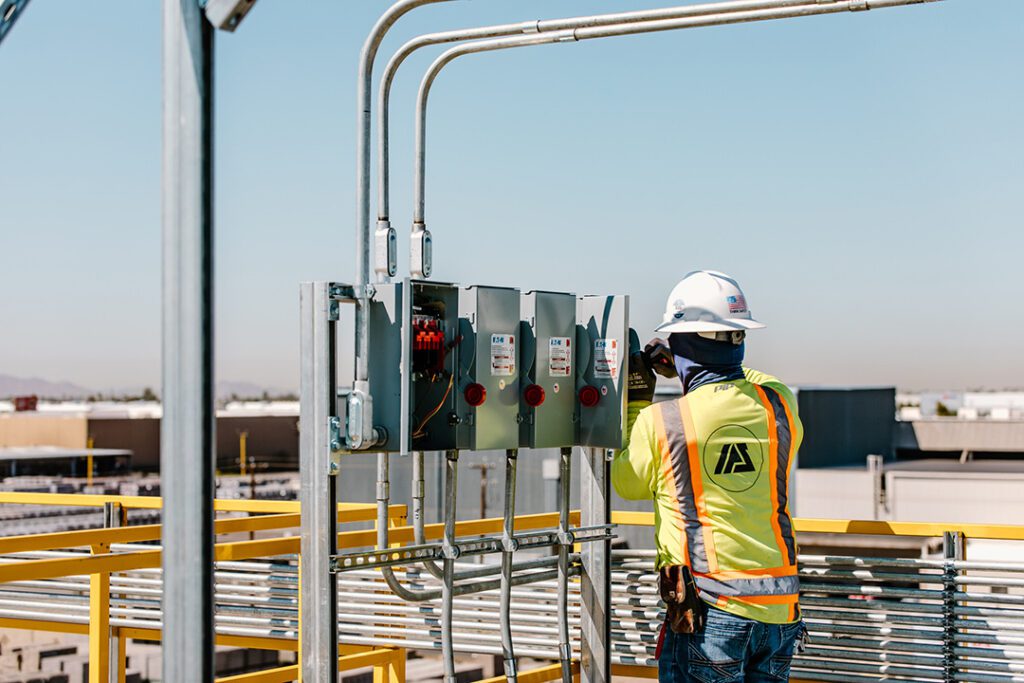 An Industrial Power Services worker in a safety vest is working on an electrical panel.